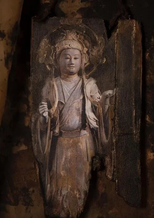 Wooden carving of a Buddhist deity or figure, with a halo around the head, holding a staff or stick in one hand, and a small object in the other, mounted on a dark wooden panel.