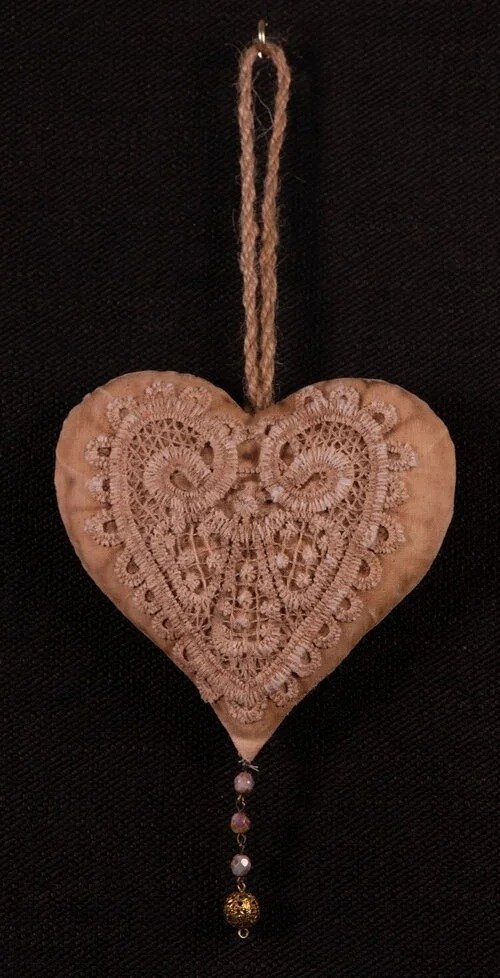 A heart-shaped ornament with intricate lace or embroidery pattern, hanging from a loop of string with a decorative bead and chain at the bottom.