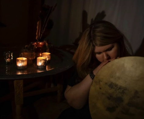 A woman with shoulder-length hair holding and kissing a large drum in a dimly lit room with candles on a table.