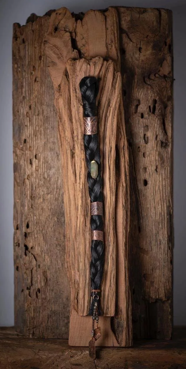 A long black braided hairpiece with copper bands and a crystal pendant, hanging vertically against a textured wooden background.