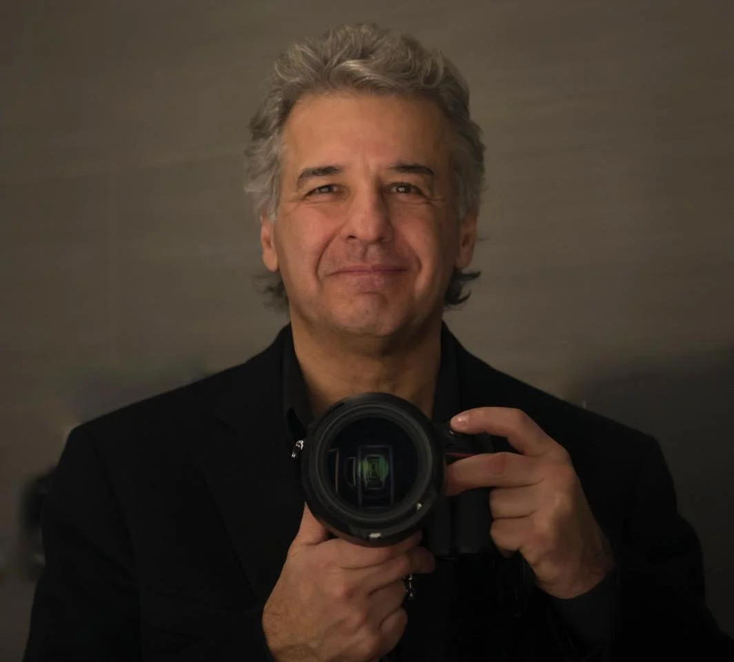 A man with graying hair holding a camera taking a selfie in front of a mirror, smiling slightly.