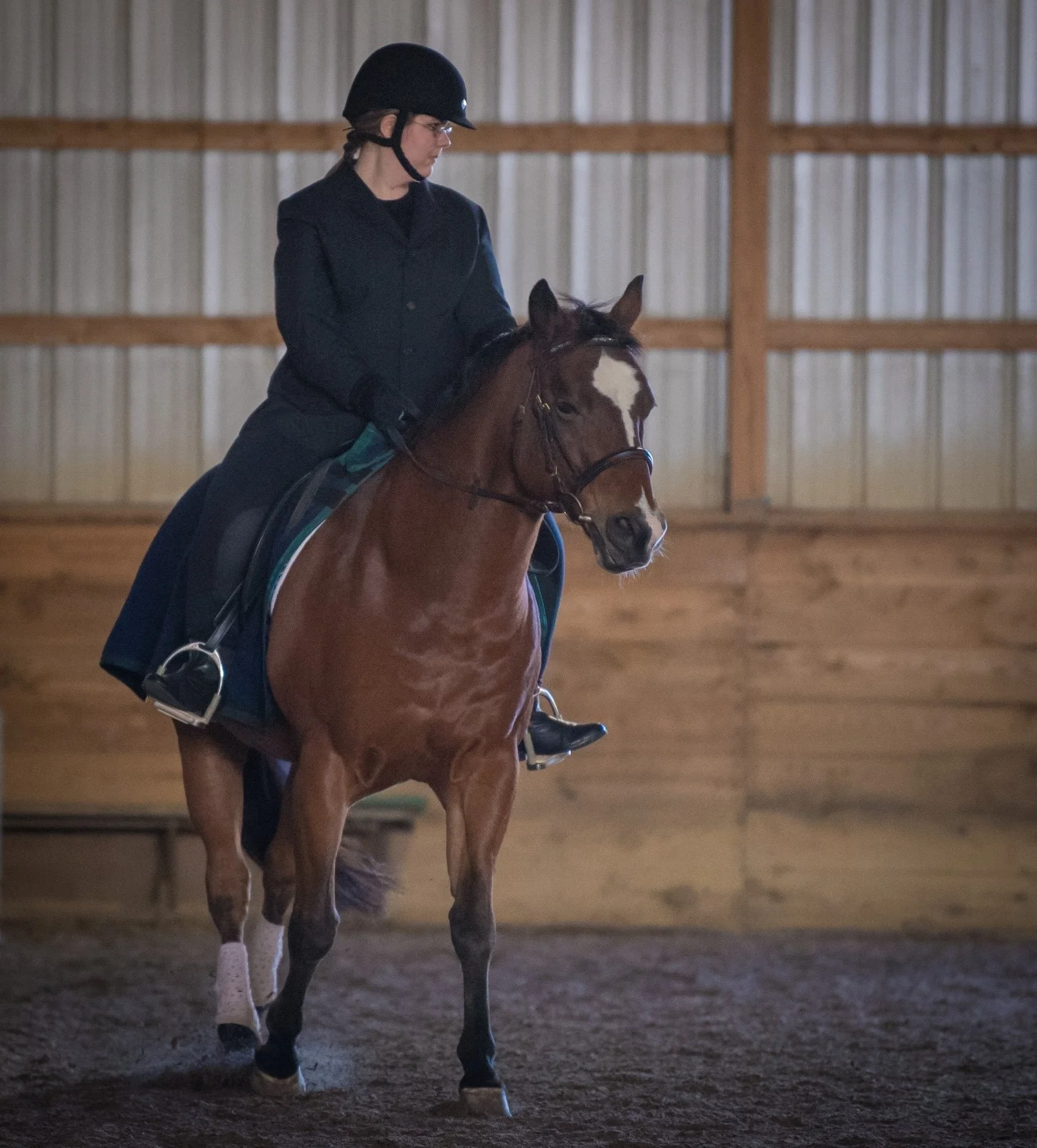 A woman wearing a black riding helmet and glasses riding a brown horse inside an indoor riding arena with wooden walls.