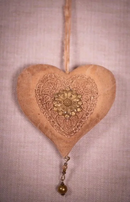 Heart-shaped decorative wooden object with gold ornate embellishment and chain with pearl hanging from the bottom, against a light fabric background.