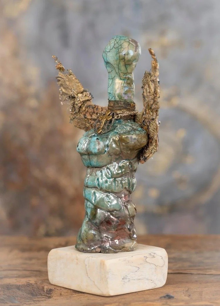 Abstract sculpture resembling a stylized human figure with wings, made of textured material with an iridescent finish, mounted on a white marble base.