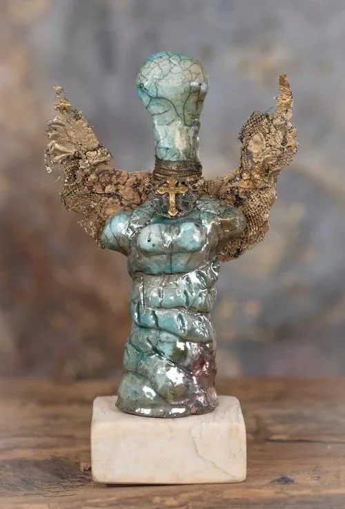 An abstract sculpture resembling a humanoid figure with a twisted, textured body, wings made of rough, earthy material, a small cross around its neck, and a bulbous head, mounted on a beige base.