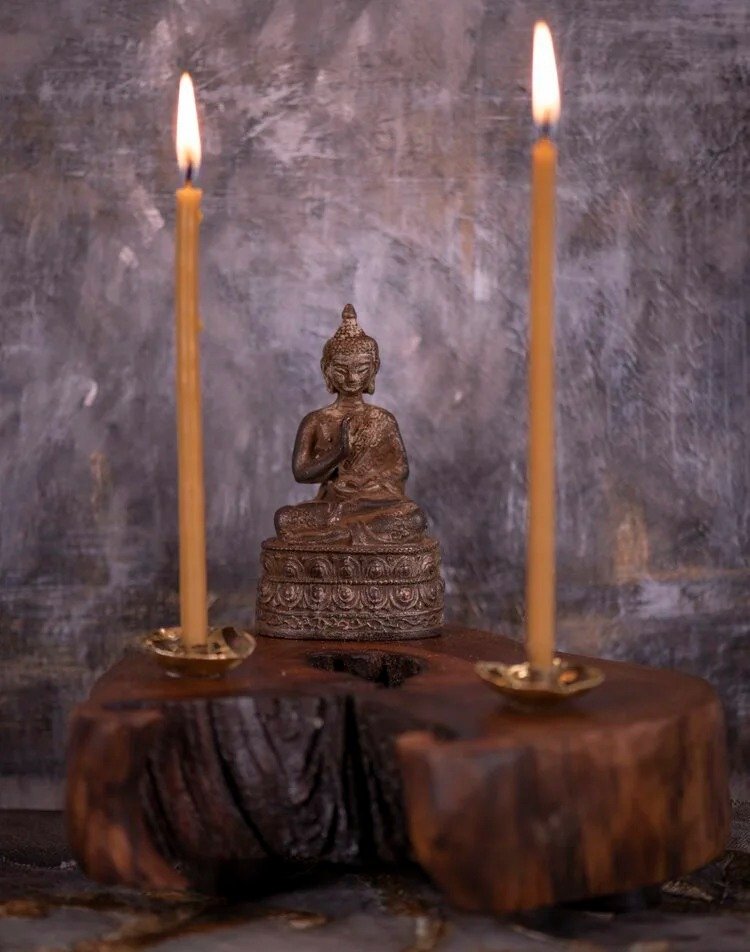 A small Buddha statue placed on a wooden surface with two lit candles on either side, set against a textured stone wall background.