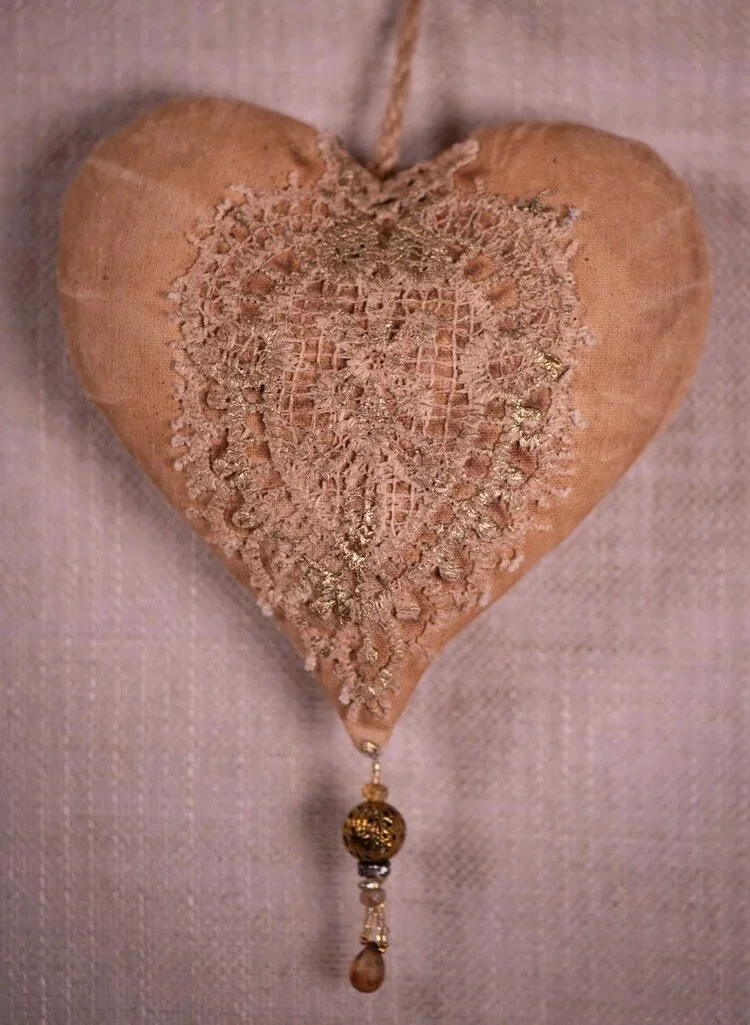 Heart-shaped fabric ornament with lace and gold embellishments, featuring a hanging bead and pearl at the bottom.