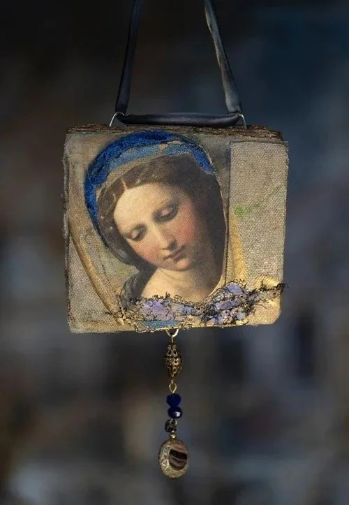 A small, square, weathered fabric-backed icon featuring a portrait of a woman with a serene expression and a dark head covering, decorated with beads and a small ornate pendant hanging below.