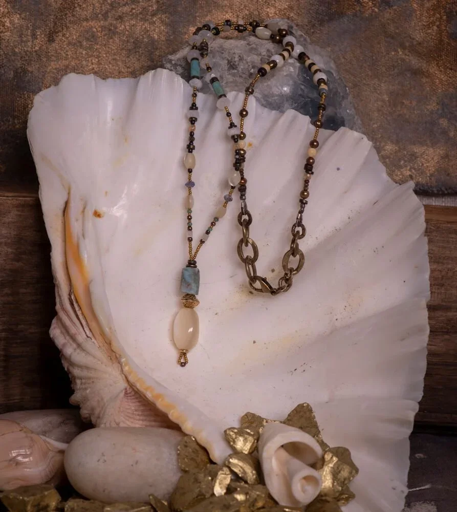 Two layered necklaces with beads and stones, draped over a large seashell, surrounded by smaller shells and gold-colored rocks.