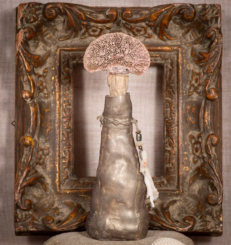 Decorative bottle sculpture with a mushroom on top, framed by an ornate, gold-accented frame with textured background.