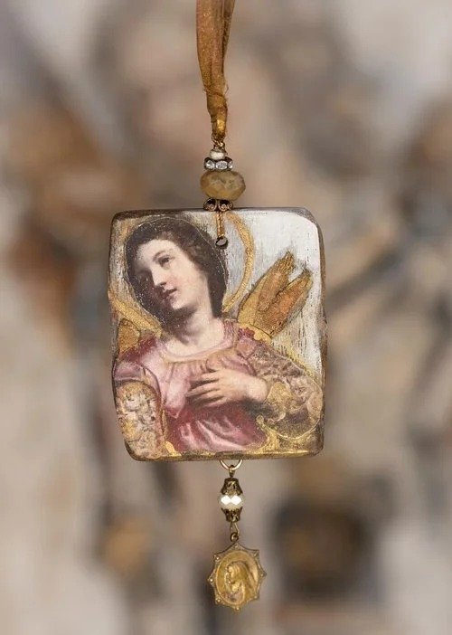 A religious pendant featuring an image of a saint or angel, with gold accents and a small hanging charm, suspended from a ribbon or cord.