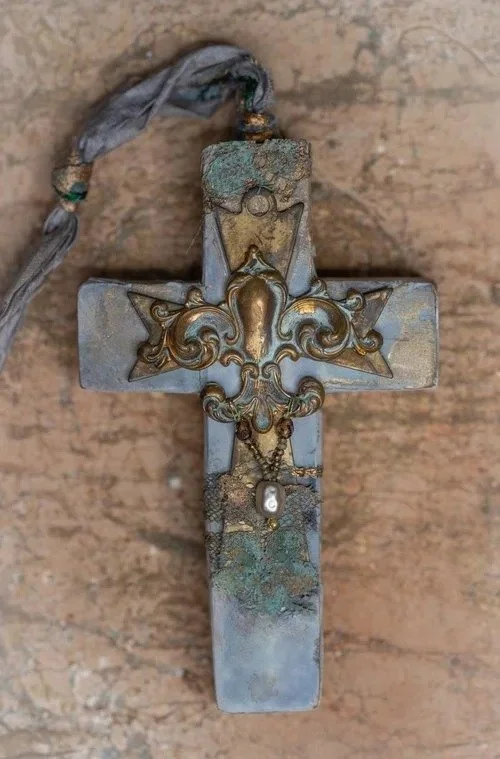 A weathered metal cross with ornate gold accents, a small pearl at the bottom, and a faded fabric in the upper left corner, resting against a wooden surface.
