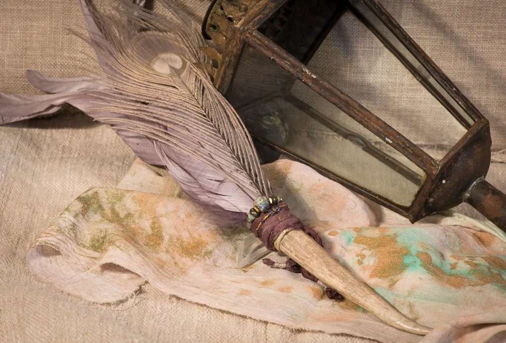 A vintage lantern, a feathered pen with beads, and a piece of fabric on a textured beige surface.