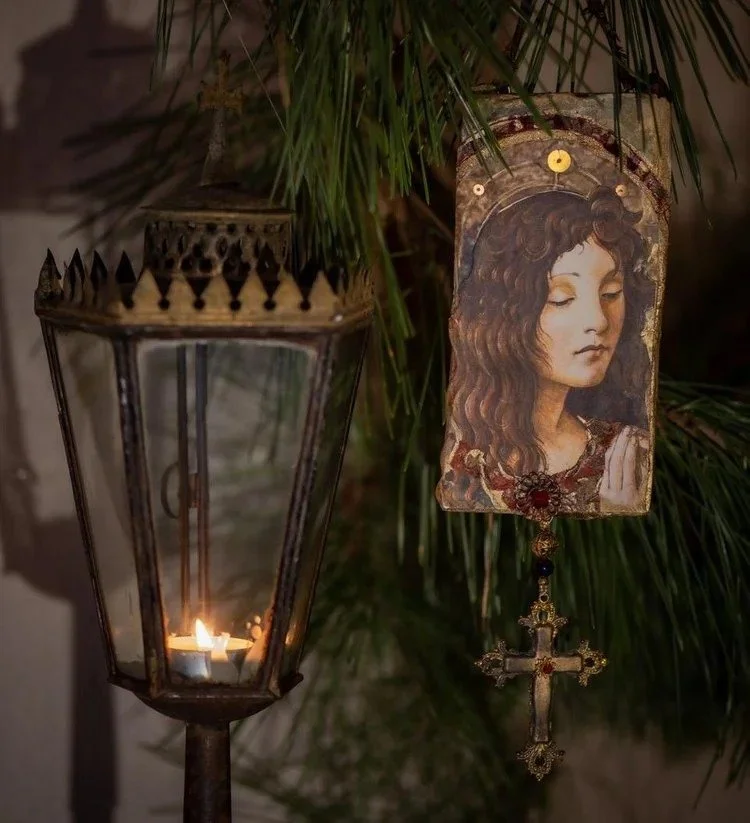 A lit candle inside a metal lantern and a religious icon depicting a woman in prayer, hanging from a green plant branch.