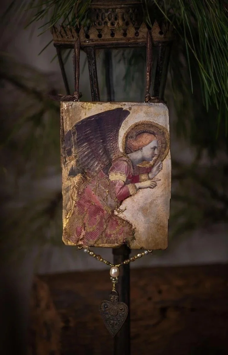 A religious icon depicting an angel with large wings and a halo, dressed in a gold and red garment, held or displayed on a stand with decorative beads and a small heart-shaped pendant hanging below. The background includes green foliage.