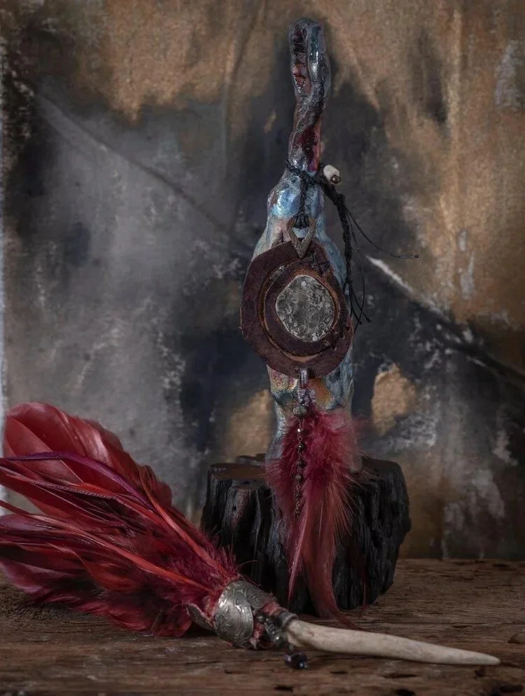 A wooden sculpture of a bird with a long neck and open beak, decorated with a circular mirror, feathers, and leather accessories, set against a rock background.