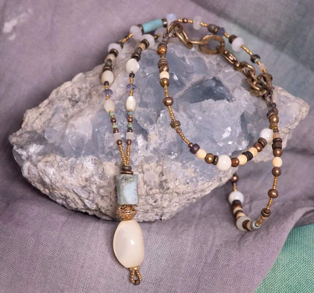 Beaded necklace with white, brown, and blue beads, featuring a large oval pendant, laid on a stone, against a gray fabric background.