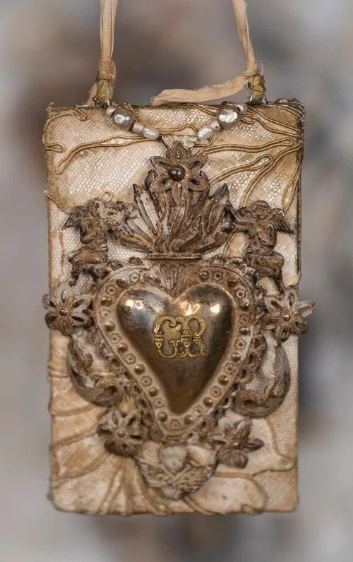 A vintage beige handbag with intricate golden and silver metallic embellishments, including a large heart-shaped ornament with a monogram, surrounded by decorative elements and angels.