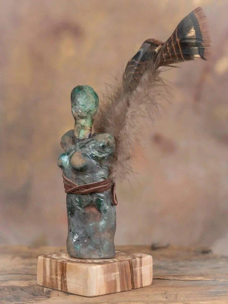 A rustic sculpture of a fallen female torso with a feather attached to the back, tied with a brown string, mounted on a wooden base.