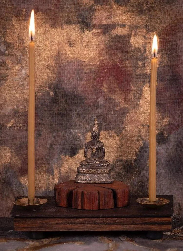 A small Buddha statue on a wooden platform, flanked by two lit candles, against a textured brown and gold background.