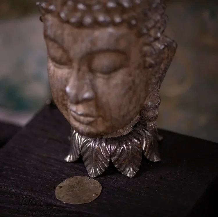 Close-up of a sculpture of a Buddha's head with closed eyes, mounted on a base with metallic leaves. There is a coin placed in front of the sculpture on a dark wooden surface.