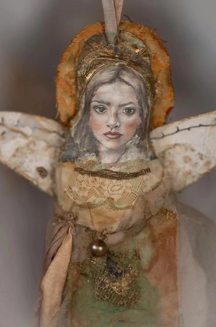 A mixed-media art piece depicting a woman's face with detailed features, integrated into a doll-like figure with fabric, lace, and decorative elements.