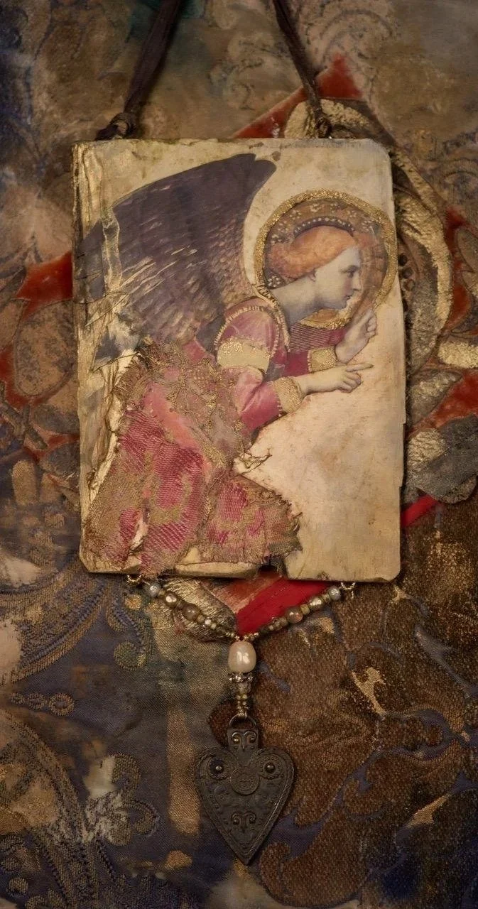 A damaged religious icon portraying an angel with a halo, pointing with one finger. The icon appears old, with torn edges and faded colors, lying on a decorative fabric surface. A necklace with a heart-shaped pendant is placed below it.