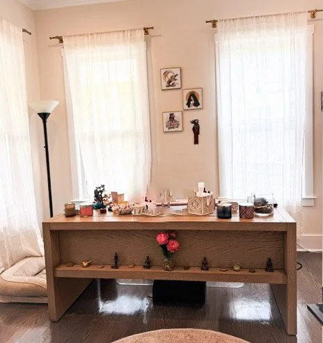 Warm and inviting room with a wooden table, pink flowers in a vase, sheer white curtains, framed artwork, and soft natural light.