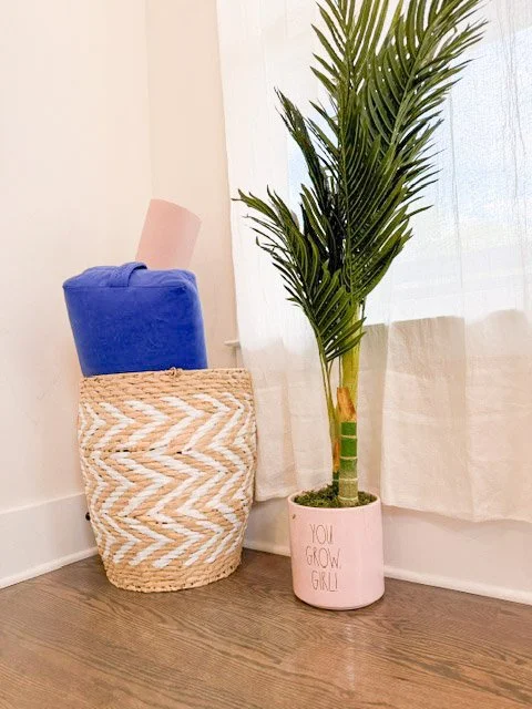 Cozy wellness space featuring a potted palm plant, rolled yoga mats, and a foam roller near a sunlit window with soft white curtains.