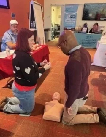A man kneeling on a pillow in front of a woman who is also kneeling, with her hands extended towards him, in what appears to be a CPR training session at an indoor event.