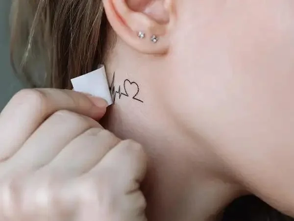 Close-up of a person's neck with a tattoo of a heartbeat line ending in a heart, and multiple earrings in the ear.