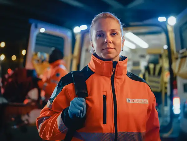 A female paramedic in an orange uniform standing in front of an ambulance at night