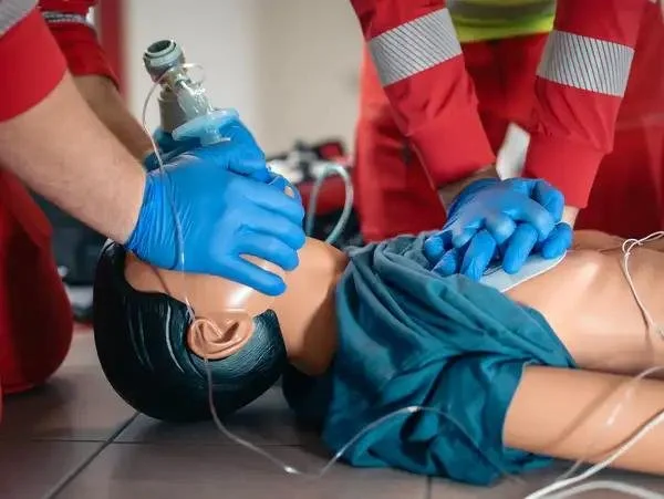 Two emergency responders in red uniforms performing CPR on a child mannequin, with one using an oxygen mask.