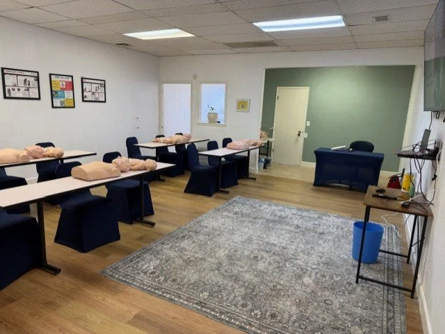 Classroom with desks, chairs, and mannequin torsos for CPR training, with a rug, window, door, and some wall decorations.