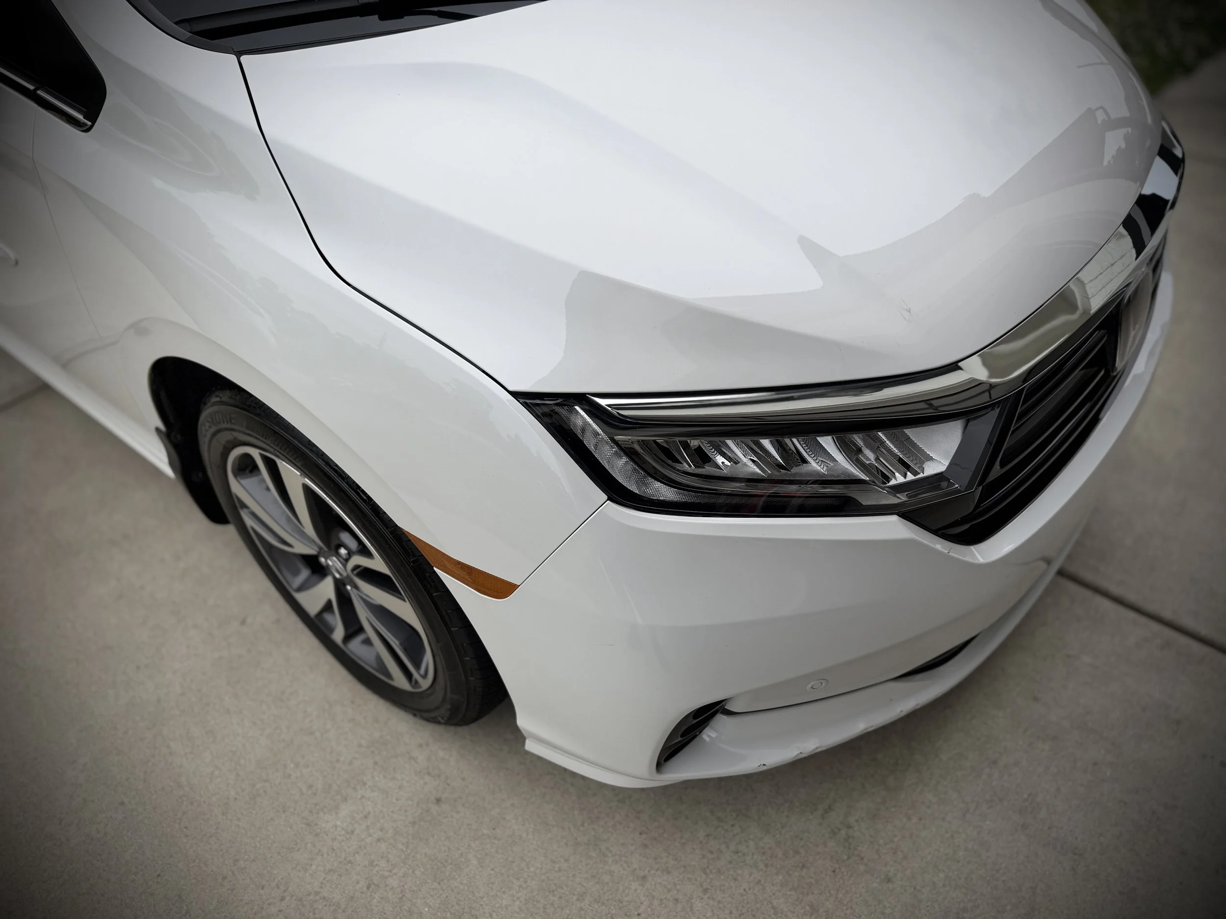 Close-up of the front part of a silver or gray modern car, showing the sleek headlight, part of the hood, front grille, and a portion of the front wheel with a stylish rim.