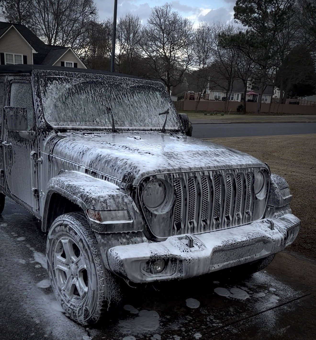 A black Jeep vehicle covered in white soap suds during washing in a driveway with trees and houses in the background.