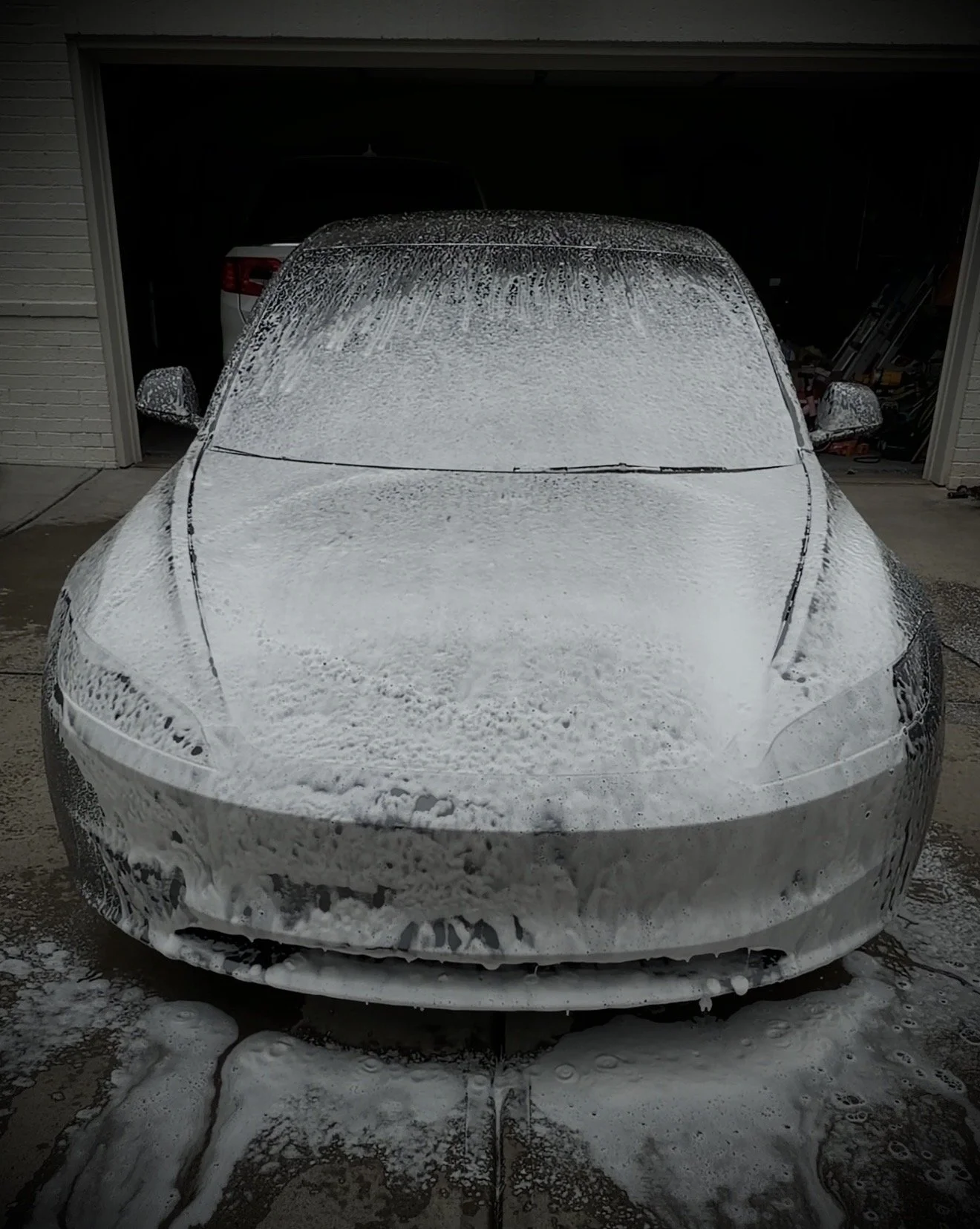 Front view of a white car covered in soap suds in a garage, with a foam bath on the ground in front.