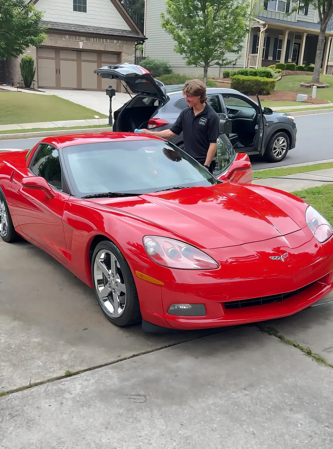 A woman is cleaning the hood of a red sports car in a residential driveway, with an open black SUV parked behind her and houses with trees in the background.
