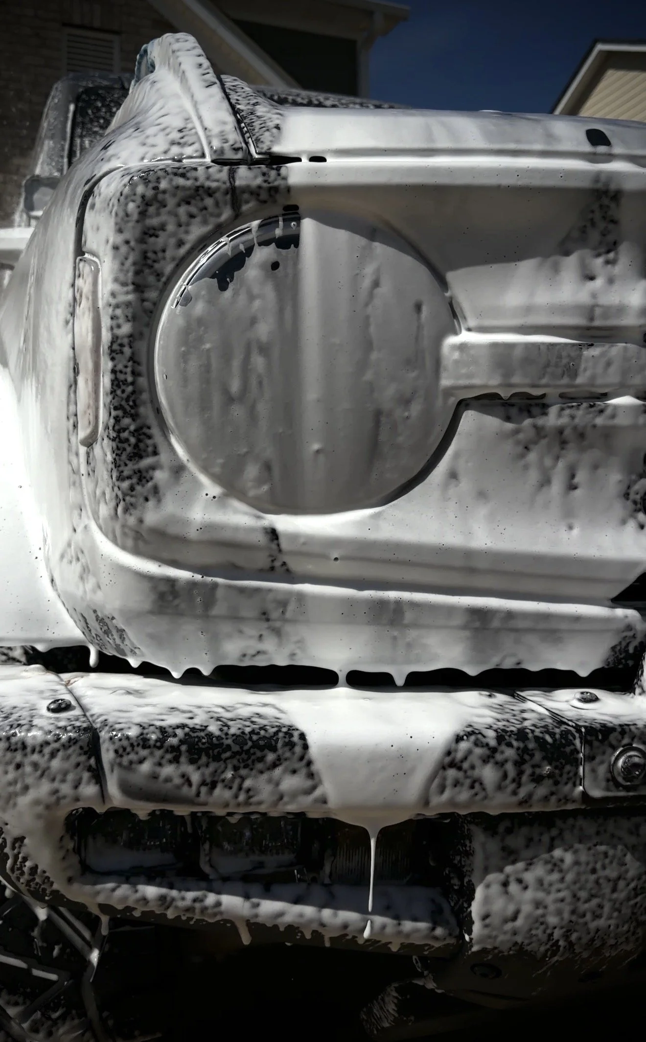 A close-up of a car's front end covered in soap suds, with a foam brush and soap visible on the vehicle and the surrounding area.