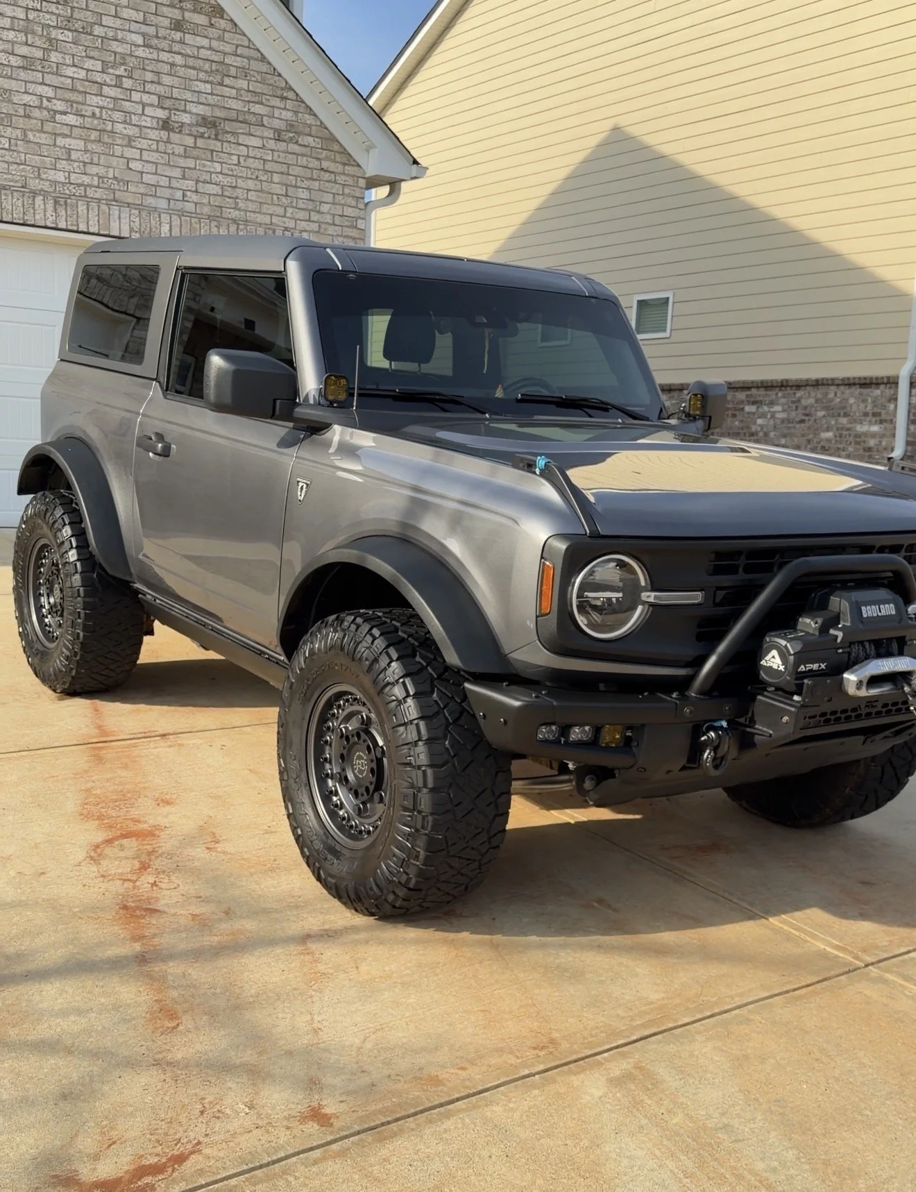A silver off-road SUV with large tires parked on a concrete driveway near a brick house with a yellow siding wall and a garage door.