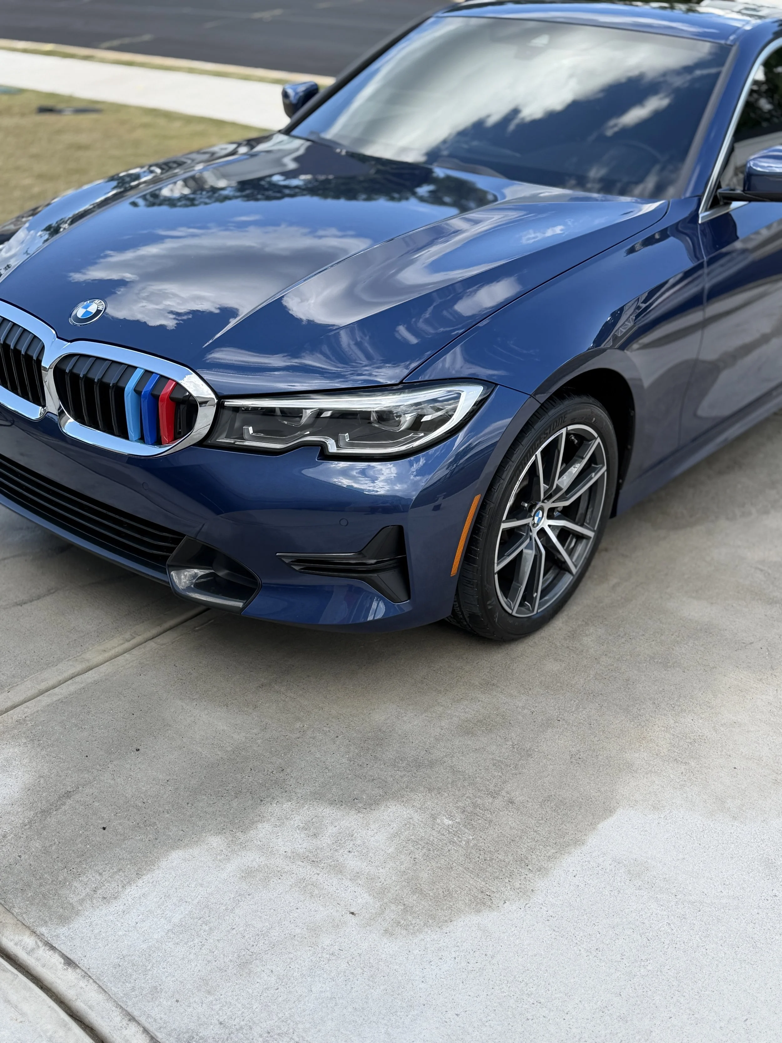 Blue BMW car parked on a concrete driveway, reflecting clouds in the shiny hood and windshield.