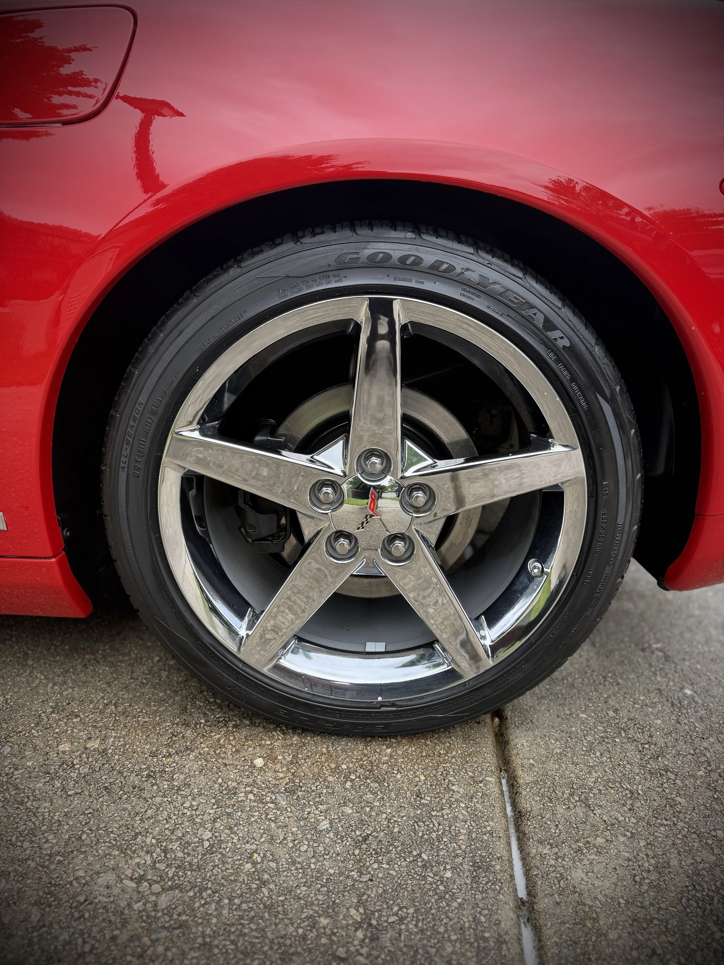 Close-up of a red sports car's front tire and wheel, with a shiny chrome rim and a red body, parked on a gravel driveway.