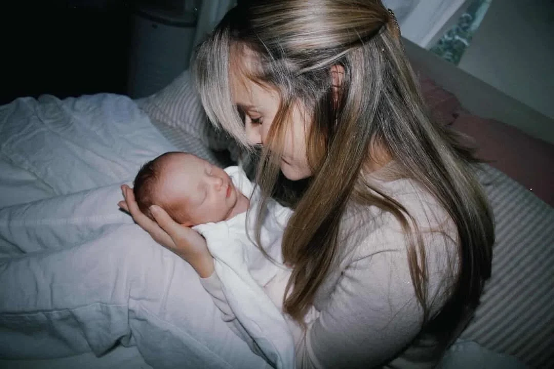 postpartum mom and sleeping newborn sitting on a bed