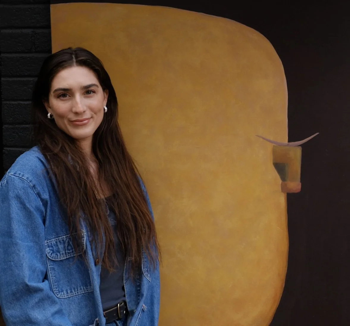 Young woman with long brown hair, wearing a blue denim jacket, standing in front of a large yellow and brown abstract painting on a black wall background.