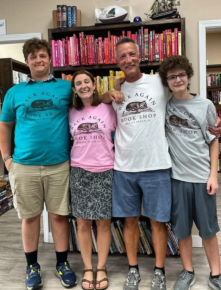A group of five people standing together inside a bookstore, smiling. They are wearing matching t-shirts that say 'Back Again Book Shop Myrtle Beach, SC.' Bookshelves filled with books are in the background.