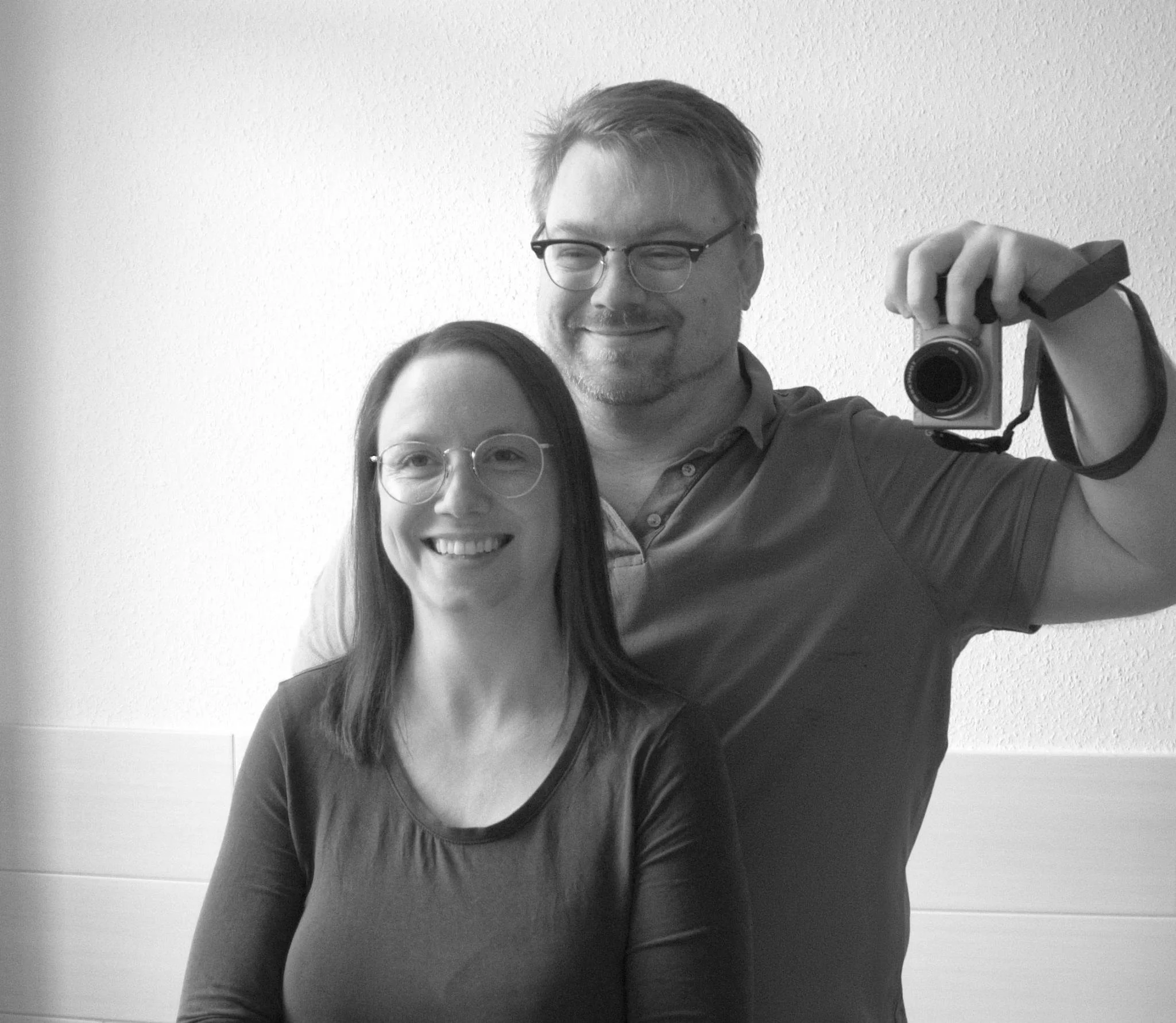 A black and white photograph of a man taking a mirror selfie. He is wearing glasses, has short hair, and is smiling. The person is holding a camera with a strap, and part of a woman's face with glasses and long hair is visible in the foreground.