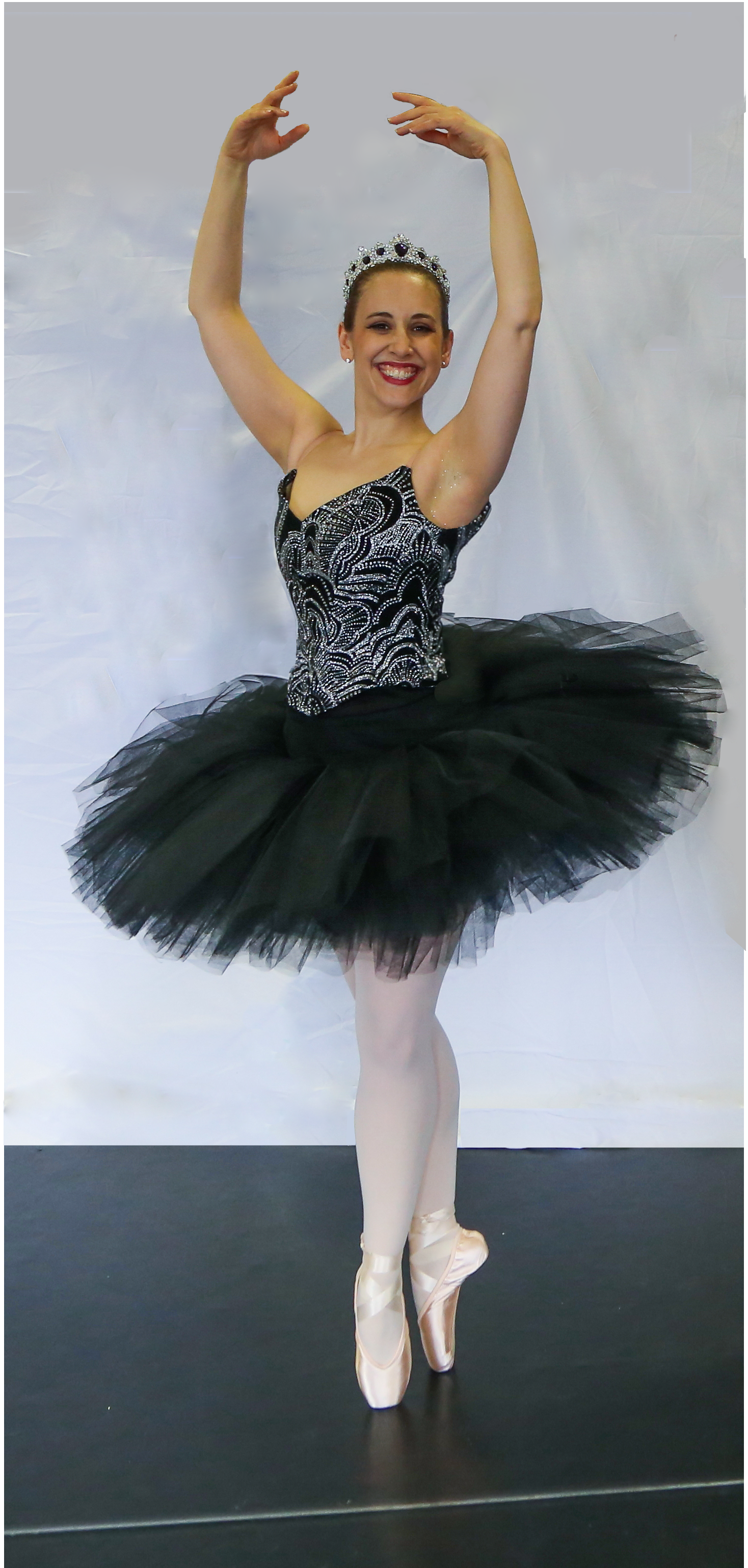 Ballet dancer in a black tutu, decorated bodice, and a tiara, standing en pointe with arms raised, smiling against a white backdrop.