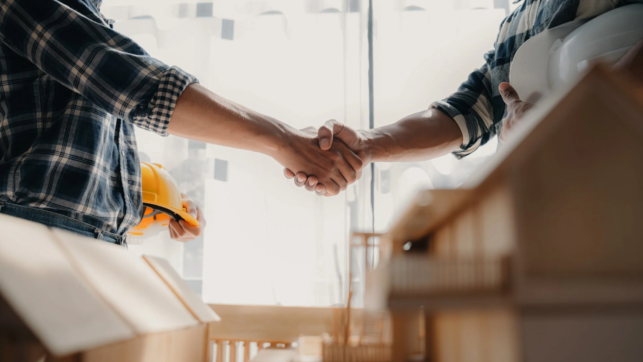 Two people shaking hands in a bright, modern office or workspace. One person is holding a yellow hard hat.