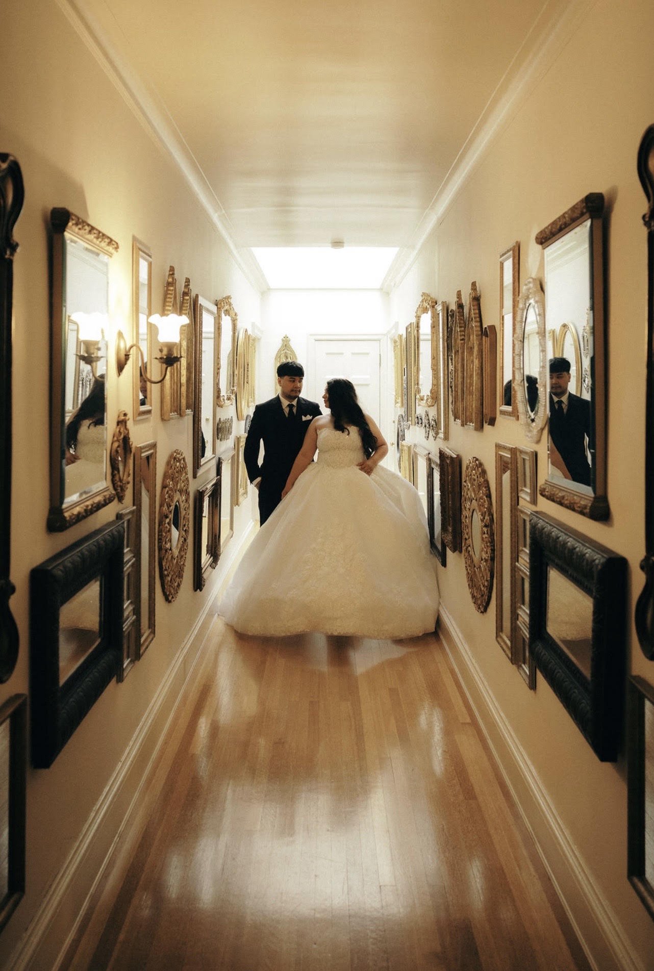 A bride and groom in wedding attire walking in a hallway decorated with framed mirrors and artwork, with warm lighting.