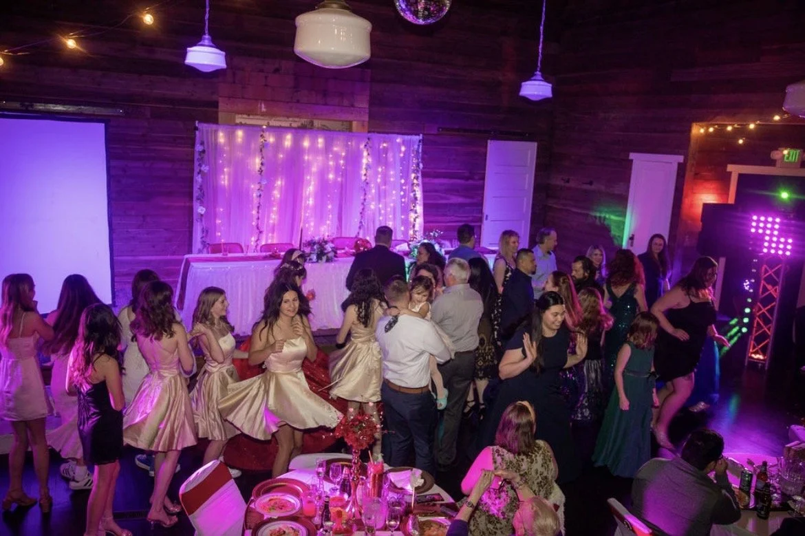 People dancing and socializing at a party with decorative lights and a dance floor, in a wooden-paneled room.